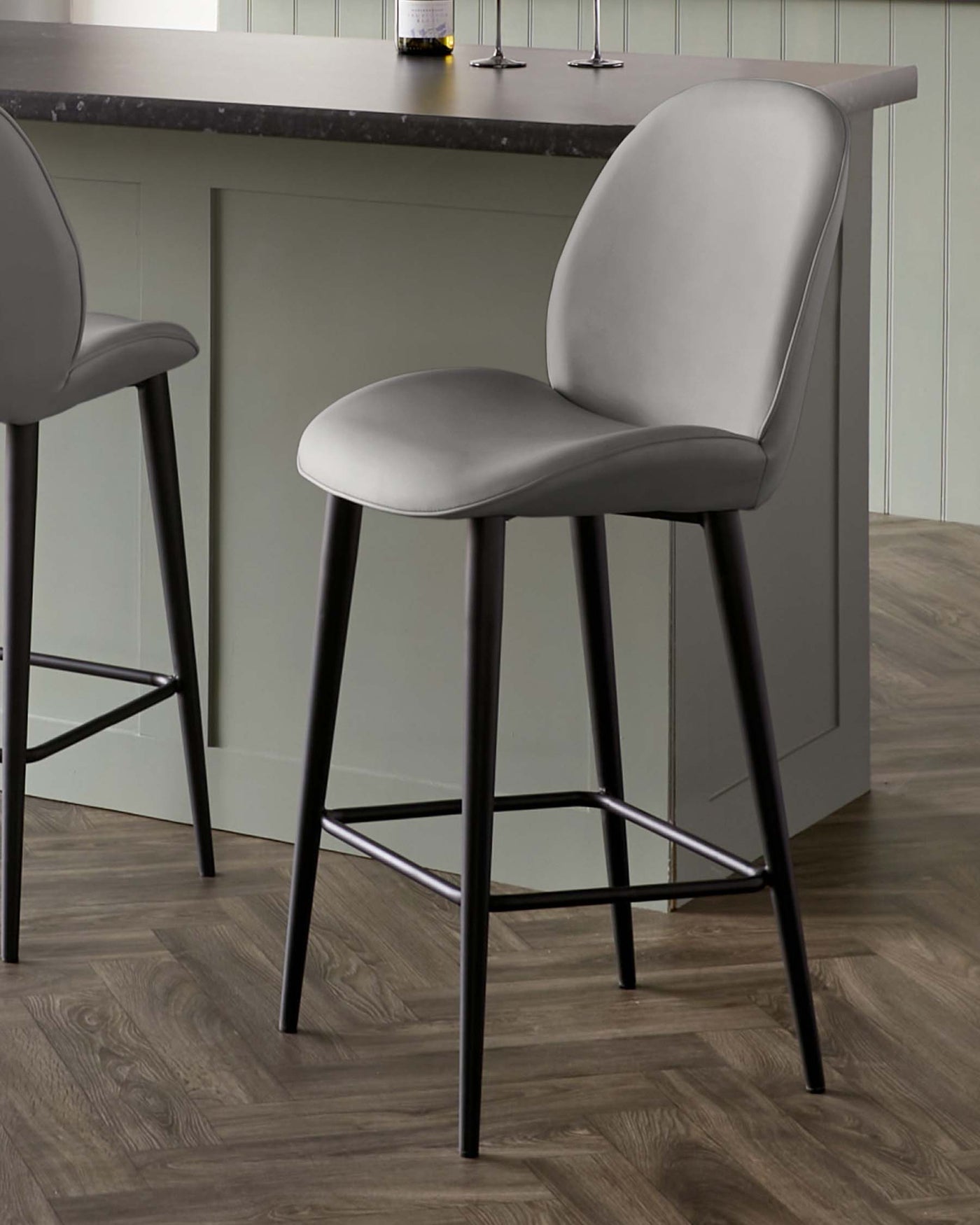 Modern barstool with a sleek grey upholstered seat and backrest on a sturdy black metal frame with a footrest bar, displayed in a contemporary kitchen setting with wooden flooring and a stone countertop.