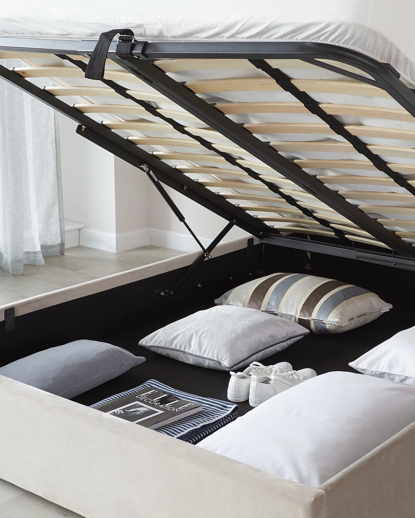 Beige upholstered storage bed with a lifted base revealing a spacious storage compartment underneath, featuring a black flooring base and a wooden slat support system.