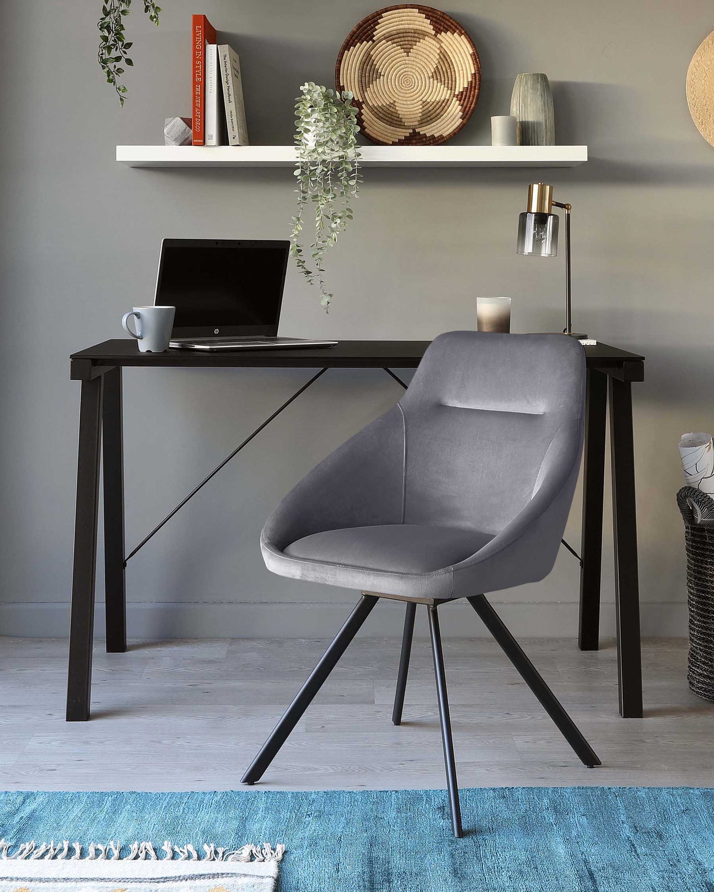 A modern home office setup featuring a sleek, black wooden desk with a subtly angled design and slender legs paired with a contemporary grey upholstered chair with a curved backrest and black metal base. The desk is accessorized with a laptop, a white mug, and a stylish desk lamp with a glass shade.