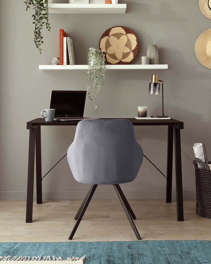 Modern minimalist home office setup with a sleek, dark wooden desk paired with a stylish grey upholstered chair with black metal legs. A white floating shelf above the desk holds decorative items and books, while a contemporary table lamp and woven basket accent the workspace. The setting is complemented by a soft blue area rug on light hardwood flooring.