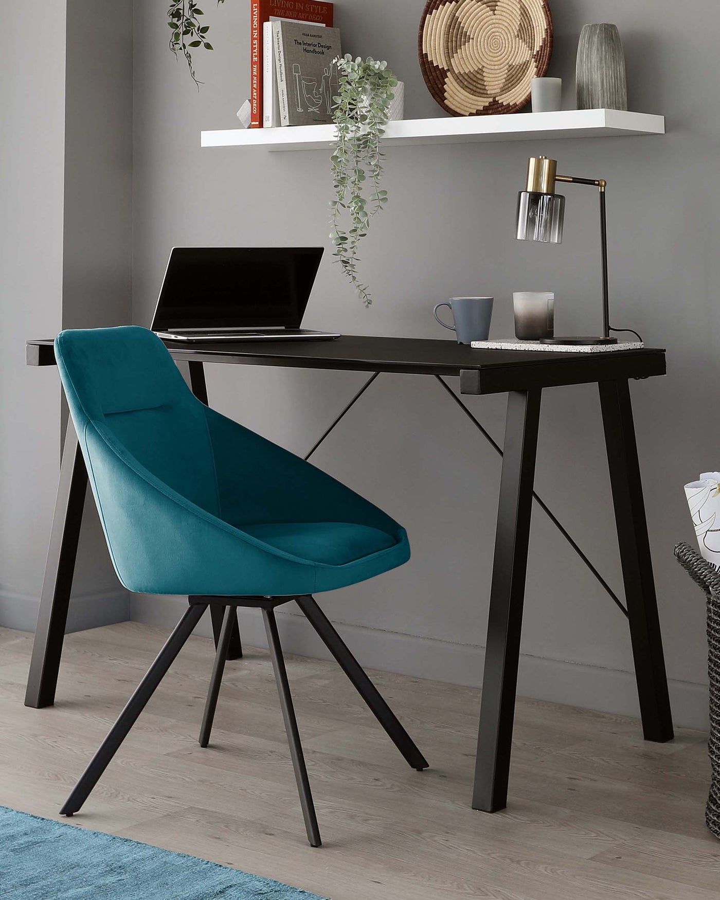A modern workspace featuring a sleek black desk with A-framed legs and a smooth surface, paired with a plush teal upholstered chair with a curved backrest and angled black legs. The desk is accessorized with an elegant brass desk lamp, a laptop, and a few cups, suggesting a functional yet stylish office setting.