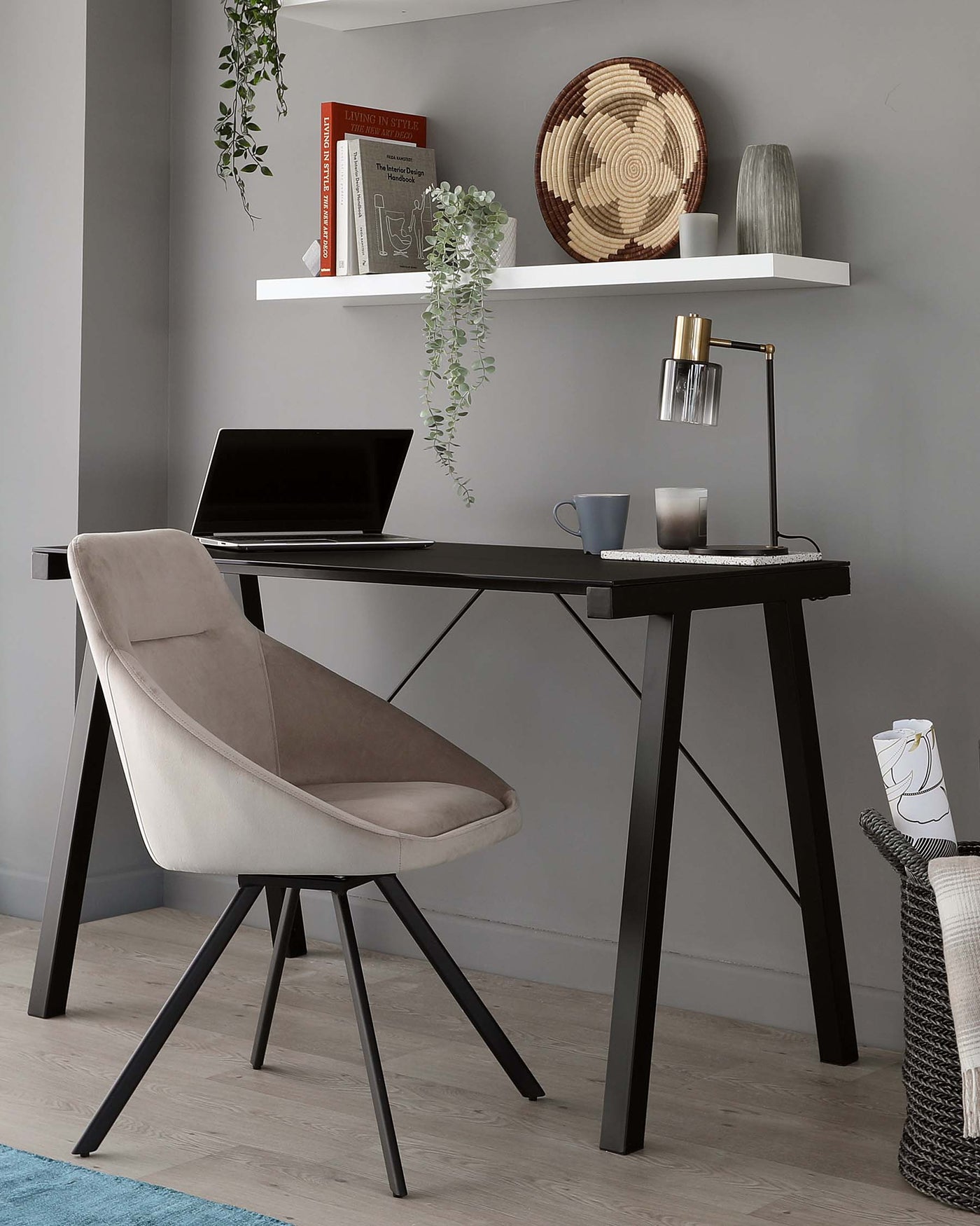 Modern home office setup featuring a sleek black desk with an A-frame leg design, paired with a plush, light taupe upholstered chair with a unique open-back and four angled legs. A white floating shelf with decorative items is mounted above.