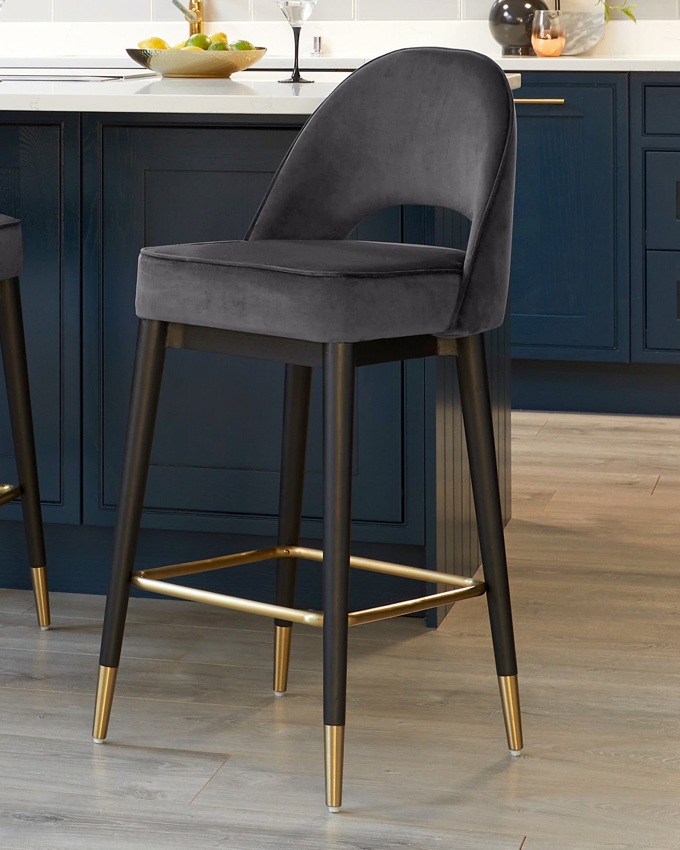 Elegant contemporary bar stool featuring a curved back, plush grey velvet upholstery, and four slender metal legs in a black finish with gold-coloured tips and a matching gold footrest.
