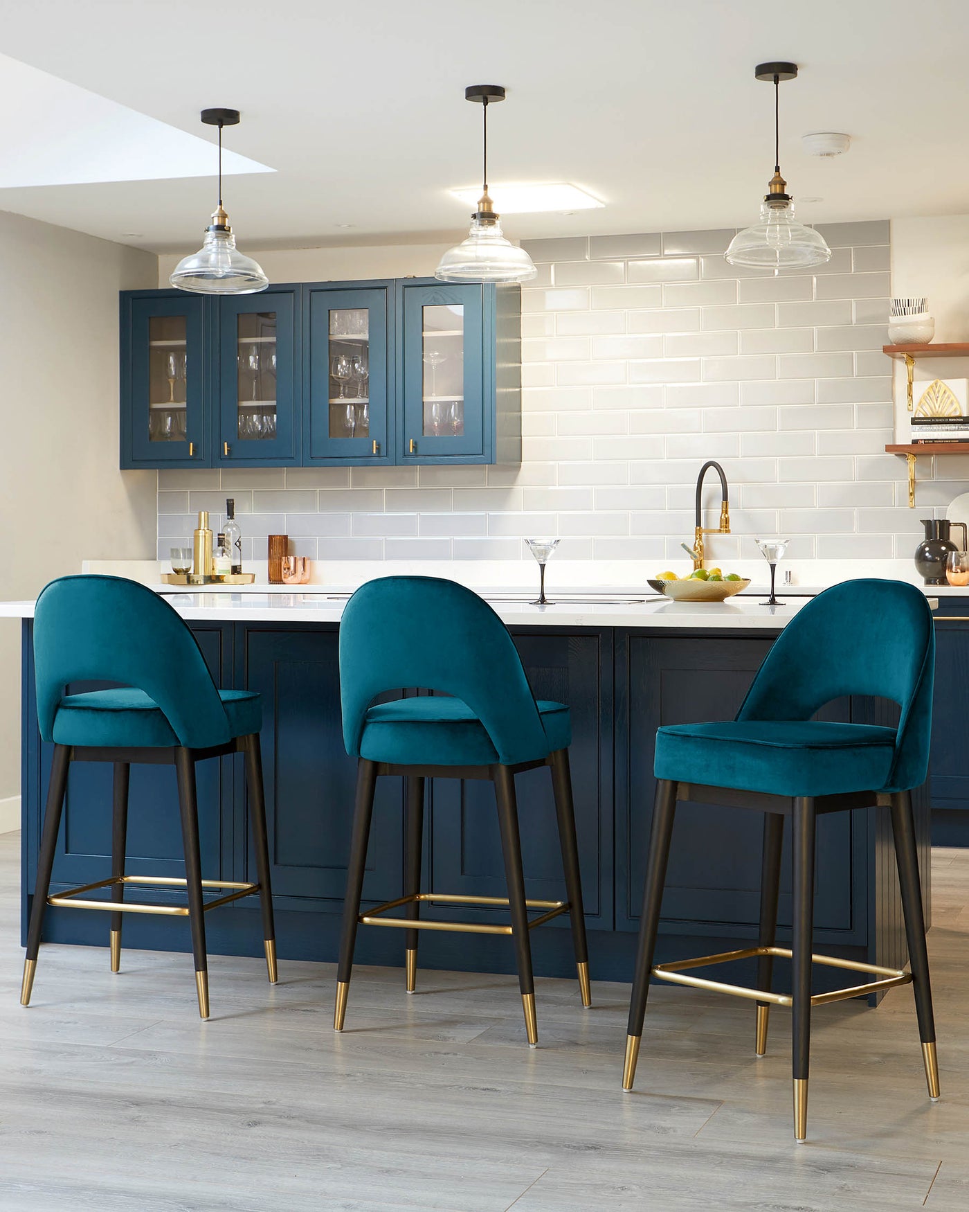 Three contemporary teal blue velvet bar stools with black legs and gold footrests in a modern kitchen setting.