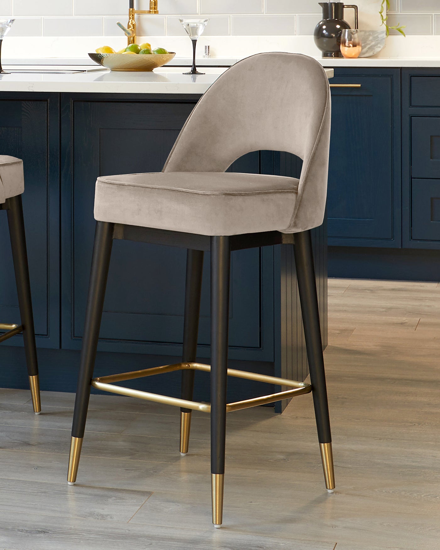 Elegant modern bar stool with a plush, curved backrest and cushioned seat in a soft grey velvet upholstery. It features a sturdy frame with matte black legs accented by metallic gold tips and footrest for added style and support. The design complements a chic, contemporary kitchen interior with dark blue cabinetry and light wood flooring.