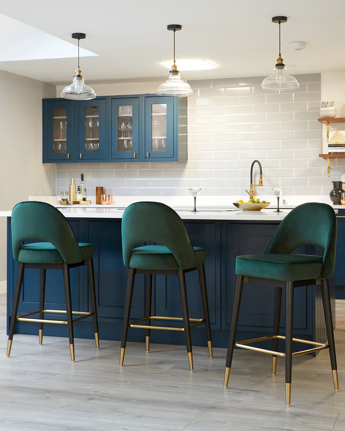 Three modern bar stools with plush dark teal upholstery and sleek black frames with gold footrests, positioned at a kitchen island with blue cabinetry.