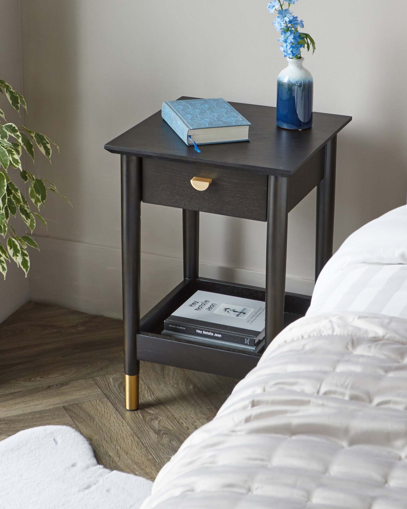 Black wooden side table with a square top, a single drawer with a brass handle, and an open lower shelf. The table stands on four legs with brass-capped feet. A blue book and a ceramic vase with blue flowers are on top of the table, and two books are placed on the lower shelf.