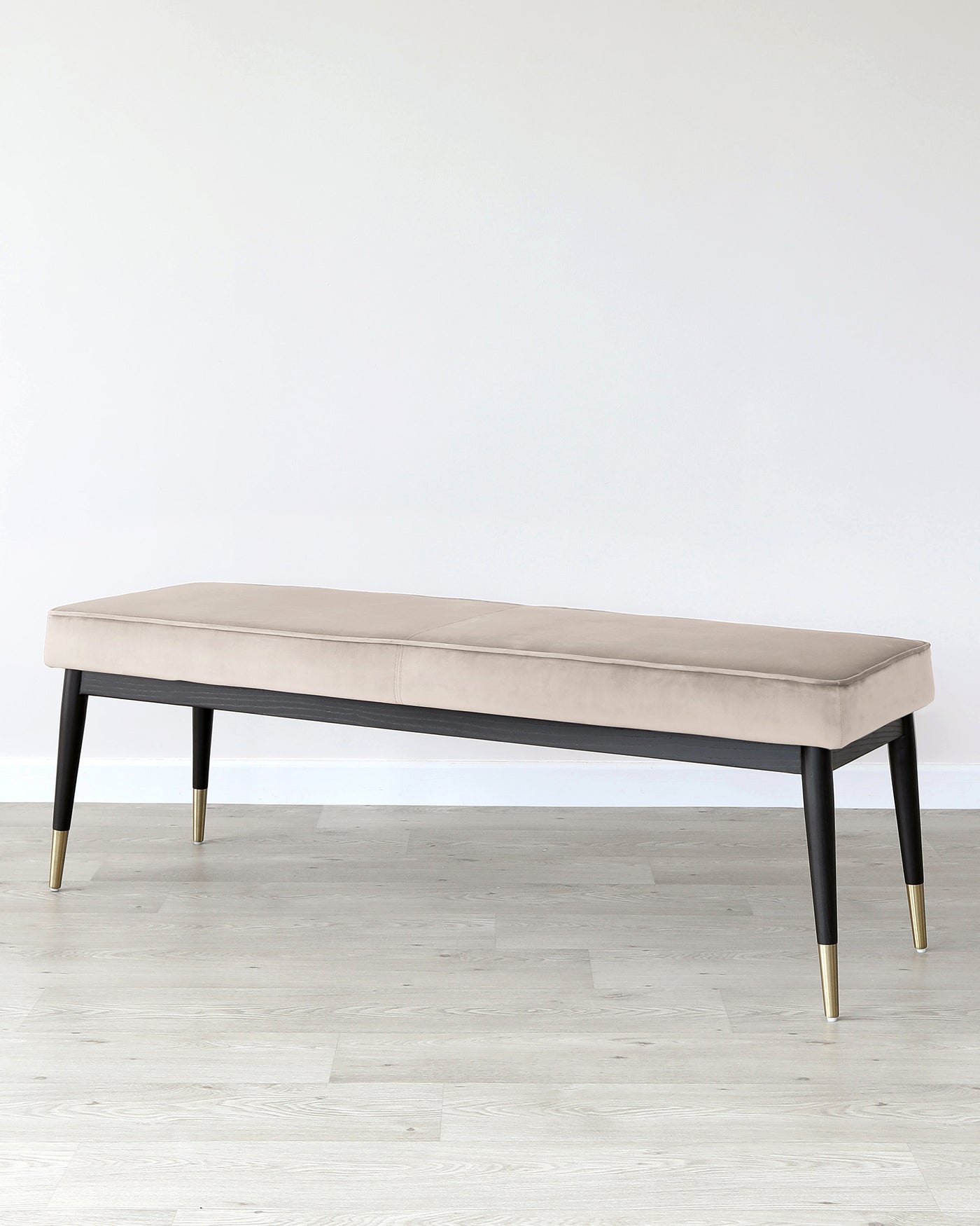 Elegant modern bench with a plush, light beige upholstered cushion on a dark wooden base with black and gold tapered legs, positioned on a light wooden floor against a white wall.