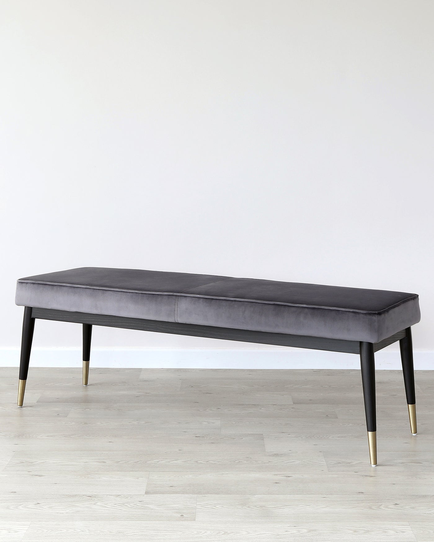 A modern upholstered bench with a sleek, grey velvet seat and black wooden legs tipped with gold metal accents. The bench stands against a neutral background, highlighting its elegant and contemporary design.