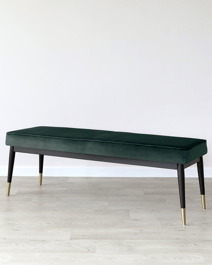 Elegant modern bench with plush green velvet upholstery and slender black legs tipped with gold caps, set against a neutral background with hardwood flooring.
