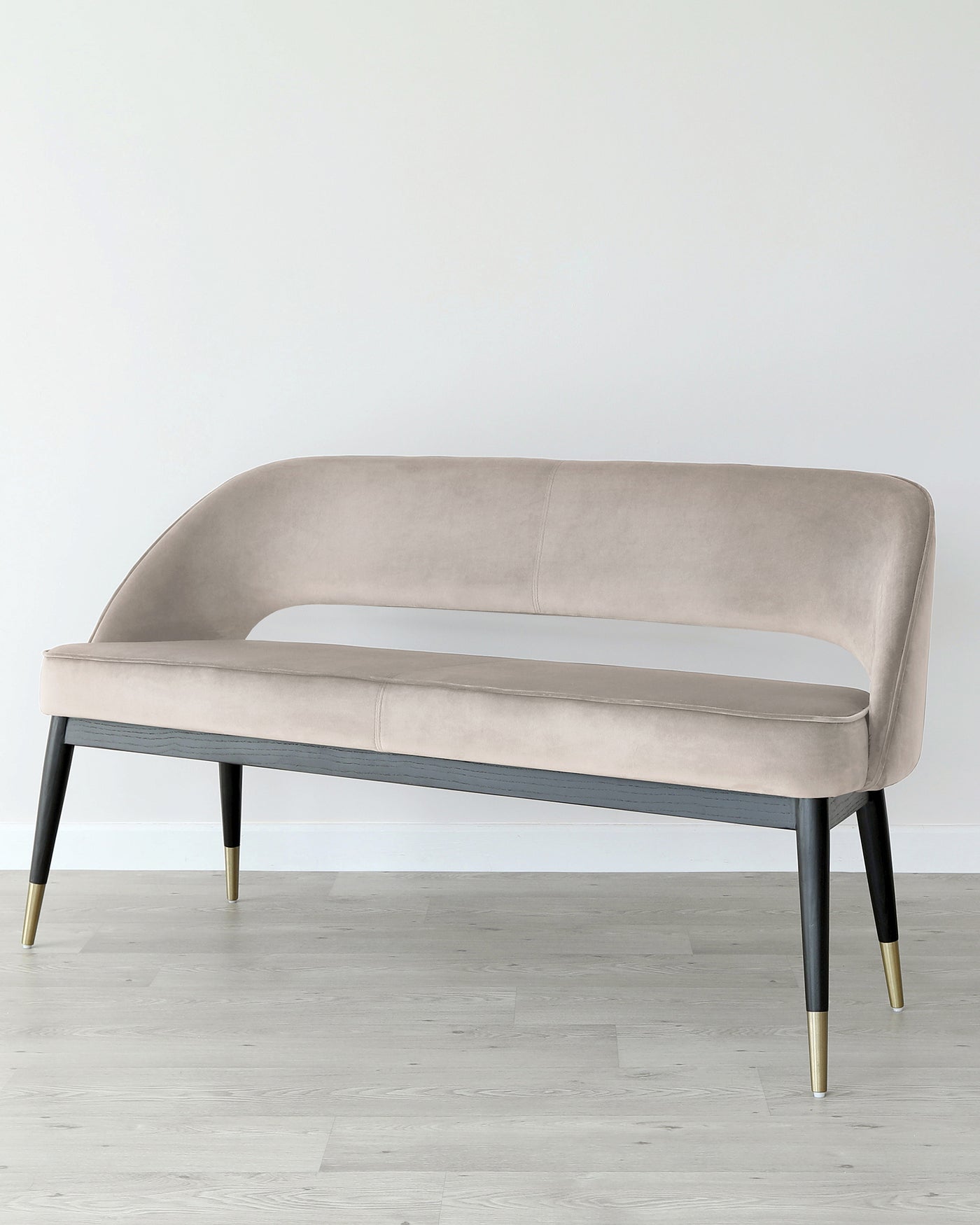 Elegant contemporary bench with a smooth, curvilinear silhouette, upholstered in a light beige fabric with a plush finish. The bench stands on four tapered wooden legs in black, accented with stylish gold-finished metal tips, set against a simple grey-floored and white-walled backdrop.