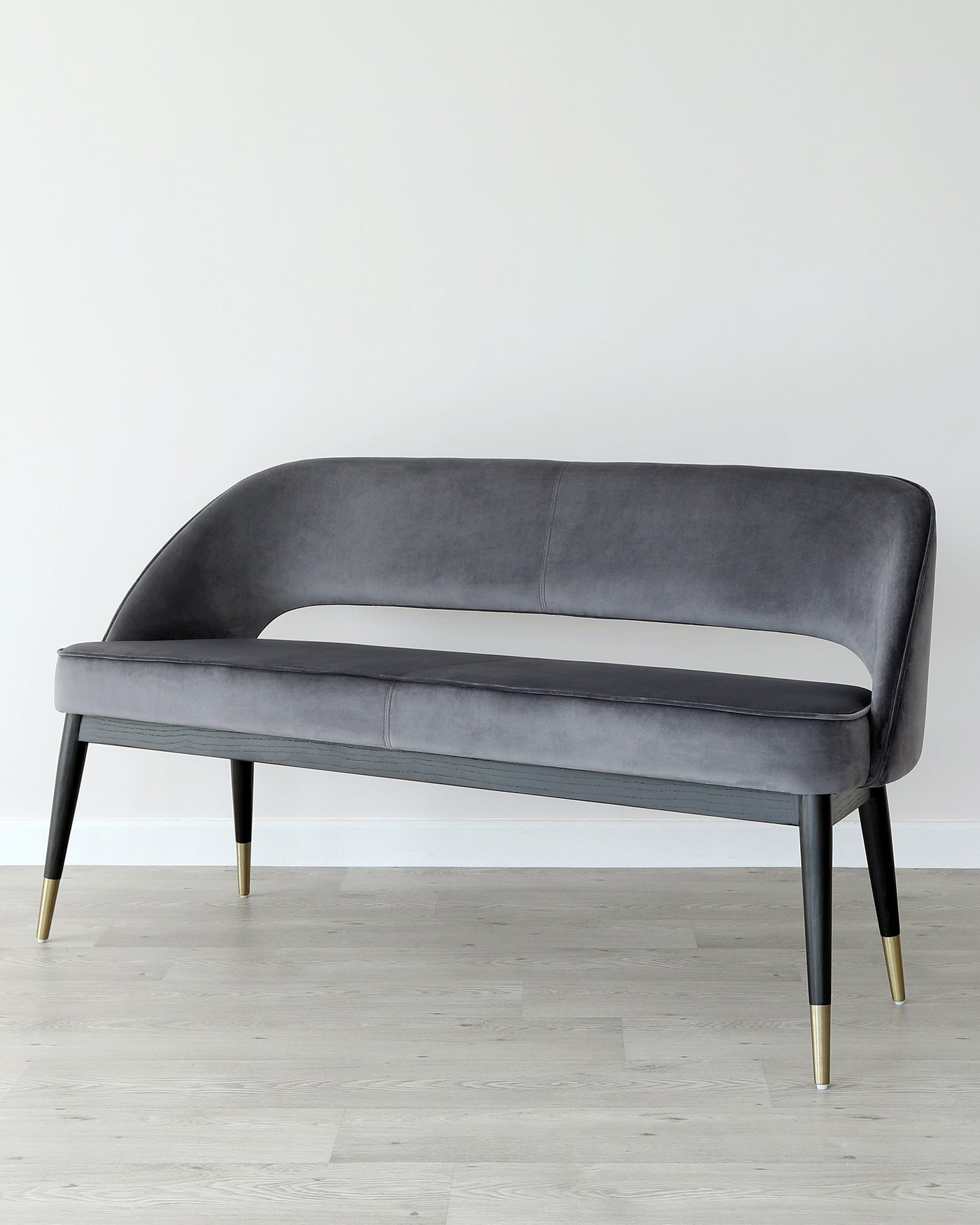Elegant modern loveseat with a curved backrest, upholstered in a smooth grey fabric, featuring sleek black legs with gold-tone metal tips, positioned on a light wooden floor against a white backdrop.
