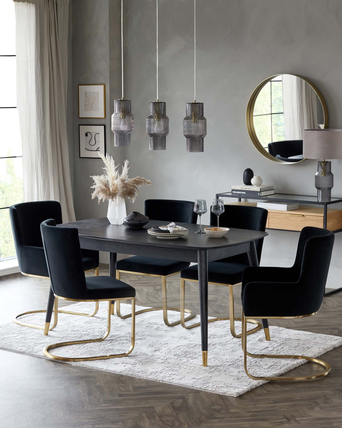 Elegant dining room set featuring a sleek rectangular black table with four plush velvet dining chairs, each with unique gold metal legs. A sideboard with a combination of black and natural wood finishes complements the table, accentuated by a circular gold-framed mirror and a stylish table lamp. A neutral-toned area rug underlines the set, and modern glass pendant lights add a sophisticated touch overhead.