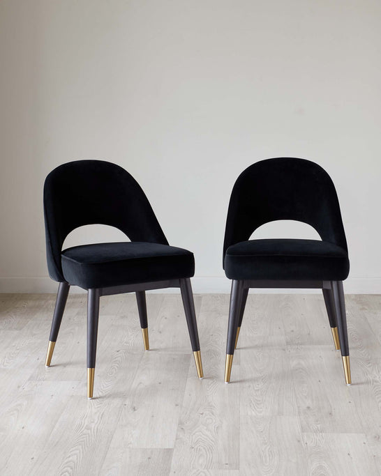 Two modern velvet dining chairs with dark upholstered seats and curved backrests, featuring sleek dark legs tipped with brass accents, positioned on a light wooden floor against a plain wall.