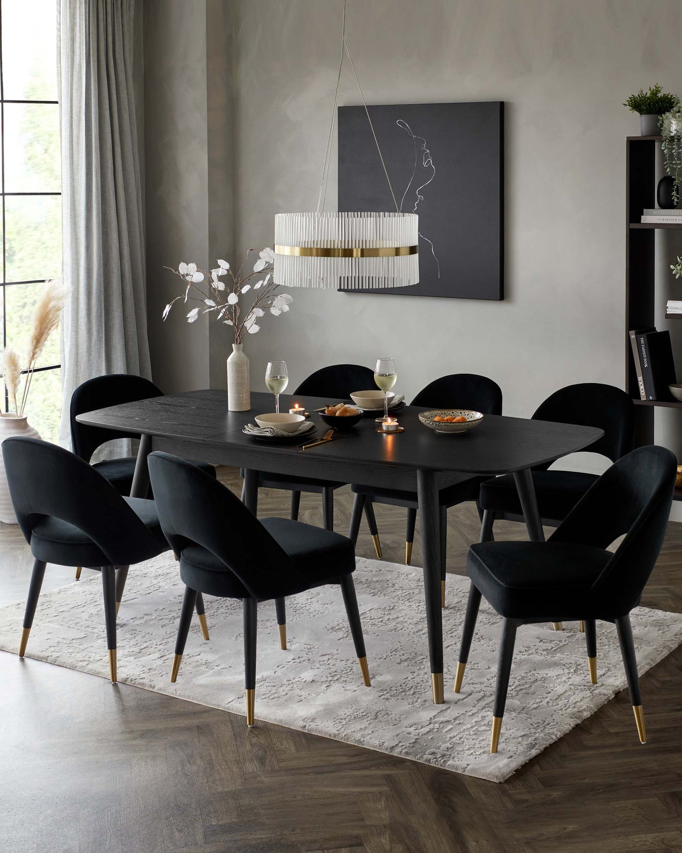 Elegant dining room featuring a modern matte black oval table with a sculptural base and brass detail. Surrounding the table are six plush black velvet chairs with curved backs and brass-tipped legs. The table is set with minimalist dinnerware, glasses, and candles, creating a sophisticated ambiance. The scene is completed by a contemporary hanging pendant light and a plush white area rug beneath the dining set.