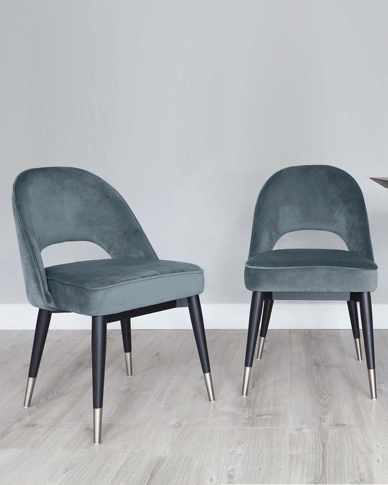 Two modern velvet dining chairs in a rich teal colour with curved backrests and cushioned seats, supported by tapered black wooden legs with metallic gold tips, set on a light wooden floor against a white wall.