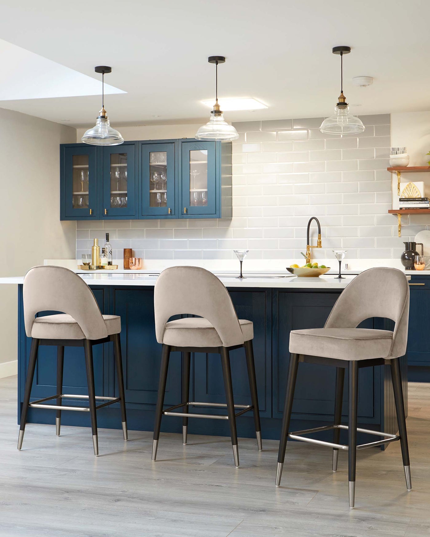 Three contemporary bar stools with grey upholstered seats and matte black metal legs are positioned at a kitchen island. The seats have curved backrests and integrated footrests on the legs for comfort. The kitchen island is dark blue with a white countertop.