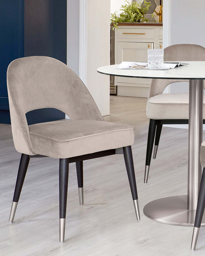 Modern dining set featuring a white round table with a sleek metal base and two plush, taupe velvet dining chairs with dark wood legs tipped with silver metallic accents.