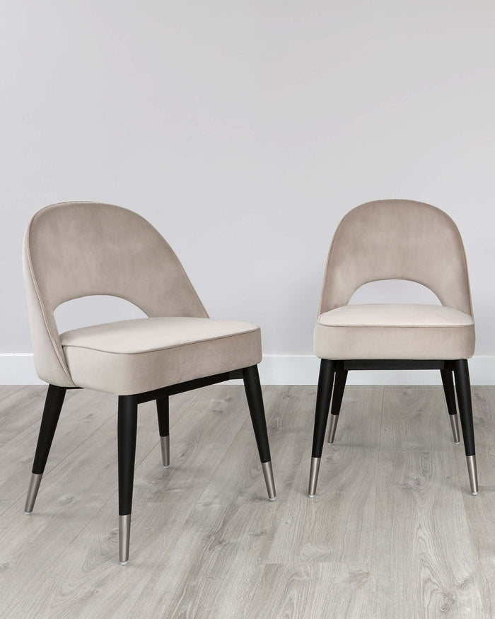 Two contemporary-style dining chairs with beige velvet upholstery and contrasting black legs with metallic tips, positioned on a grey wood-effect floor against a neutral wall.