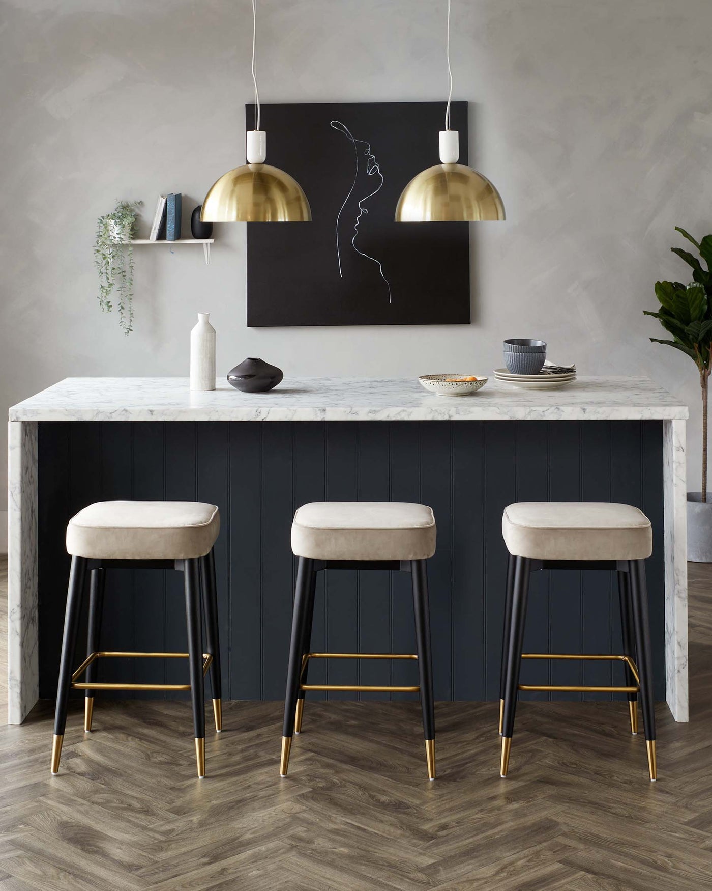Elegant modern kitchen setting featuring three bar stools with black frames and gold accents, complemented by sleek, plush, light beige seating. The stools are arranged at a kitchen island with a white marble countertop and a dark panelled base. The space is styled with minimalistic decor and soft lighting.