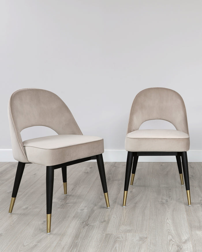 Two elegant contemporary dining chairs with beige velvet upholstery and black legs accented with gold tips, set on a light wooden floor against a white wall.