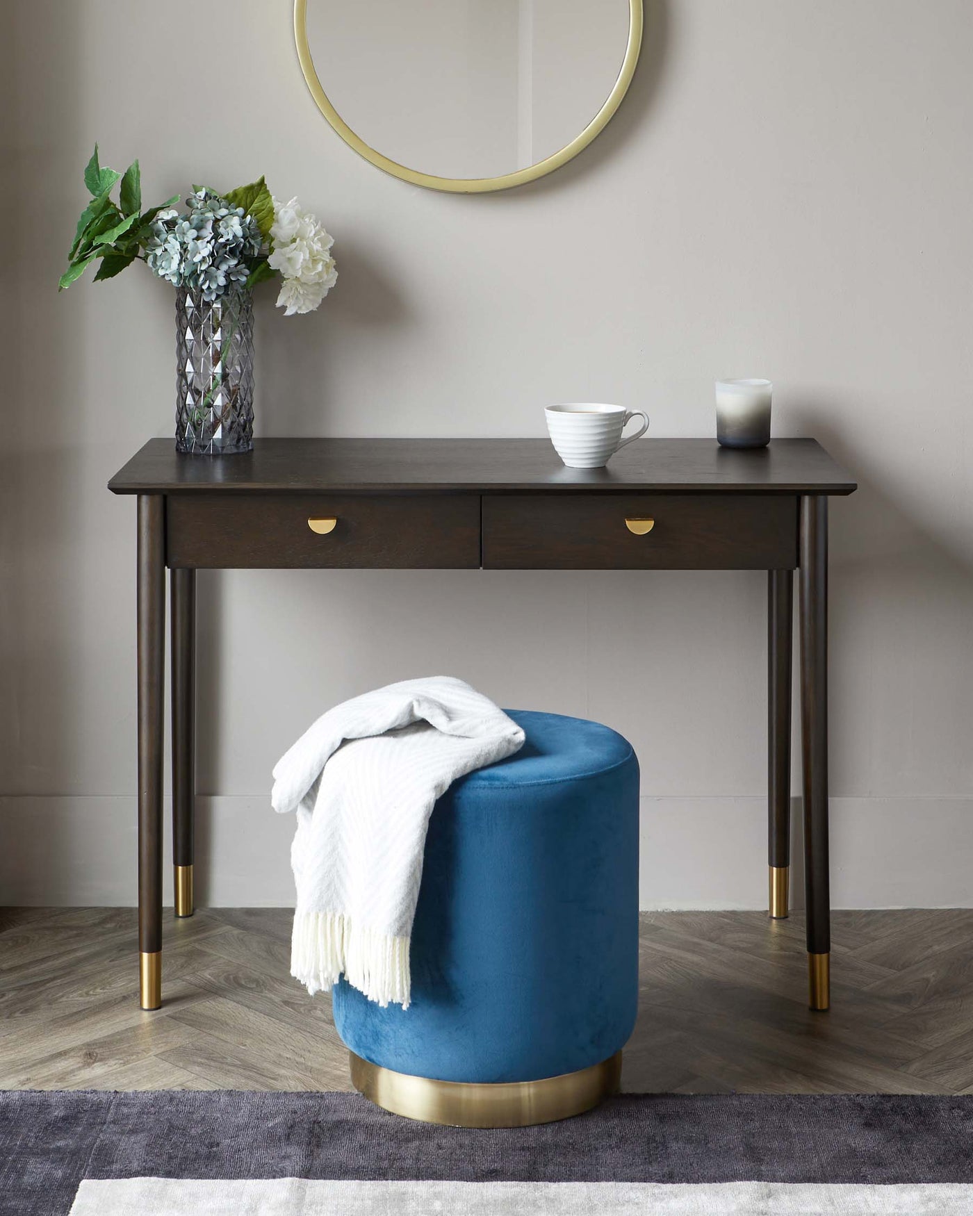 Elegant dark wood console table with two drawers and brass handles, accompanied by a plush round blue ottoman with a gold base.