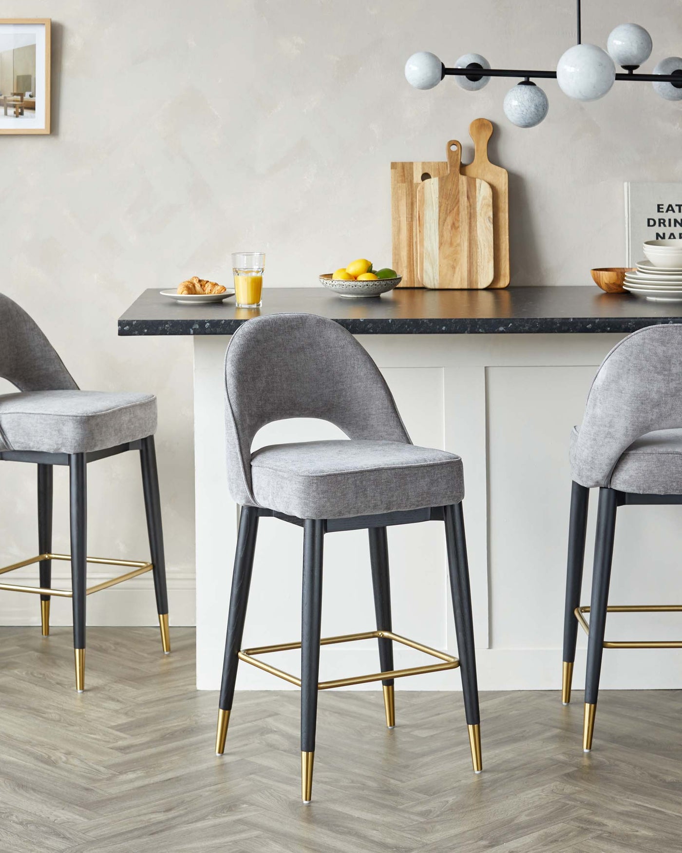 Elegant modern bar stools with grey upholstered seats and backs, black legs with gold footrests and gold accents at the bottom, positioned around a kitchen island.