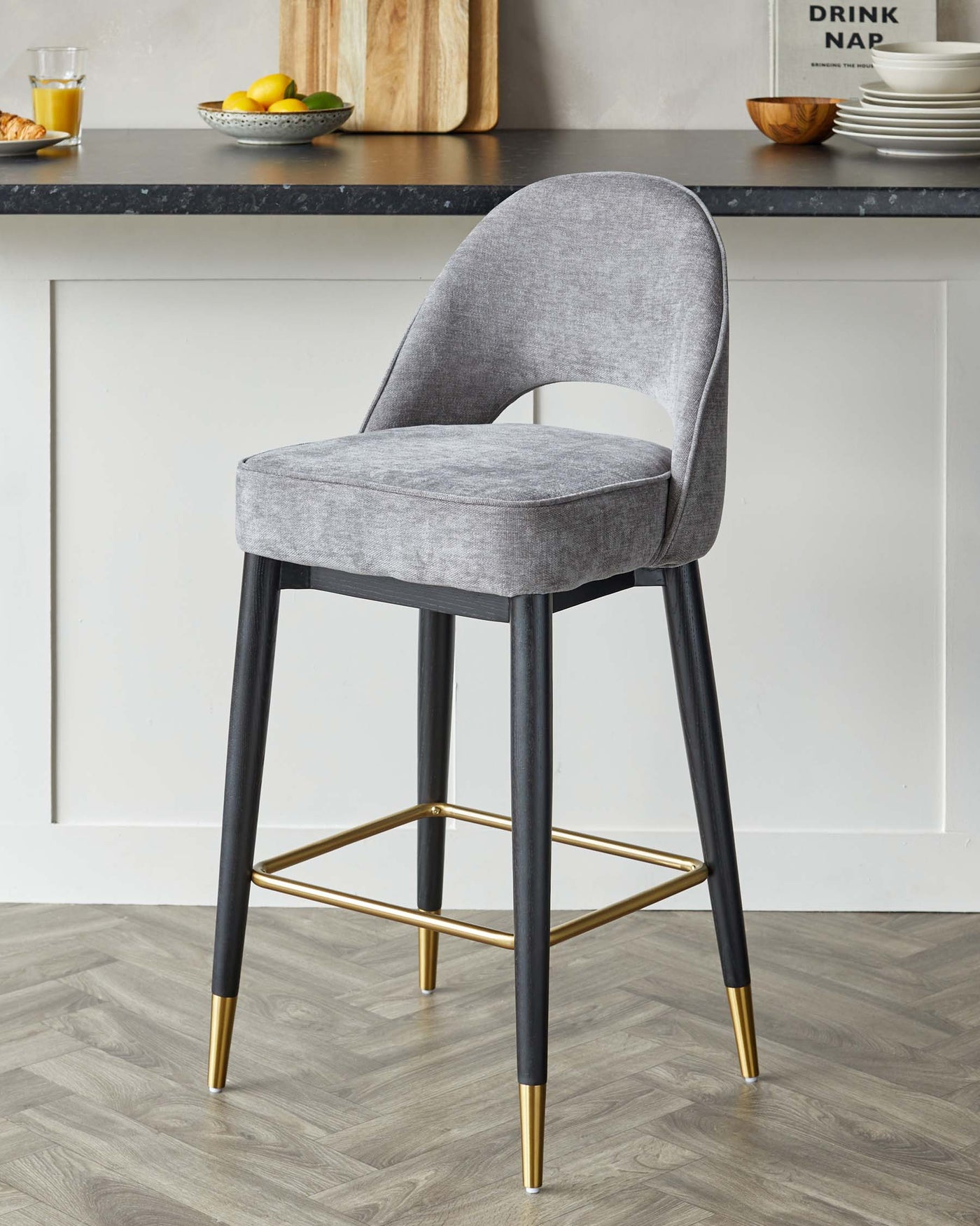 Modern grey upholstered bar stool with a curved backrest and seat on four black legs, accented with a gold footrest and gold-tipped legs, set against a kitchen counter backdrop.