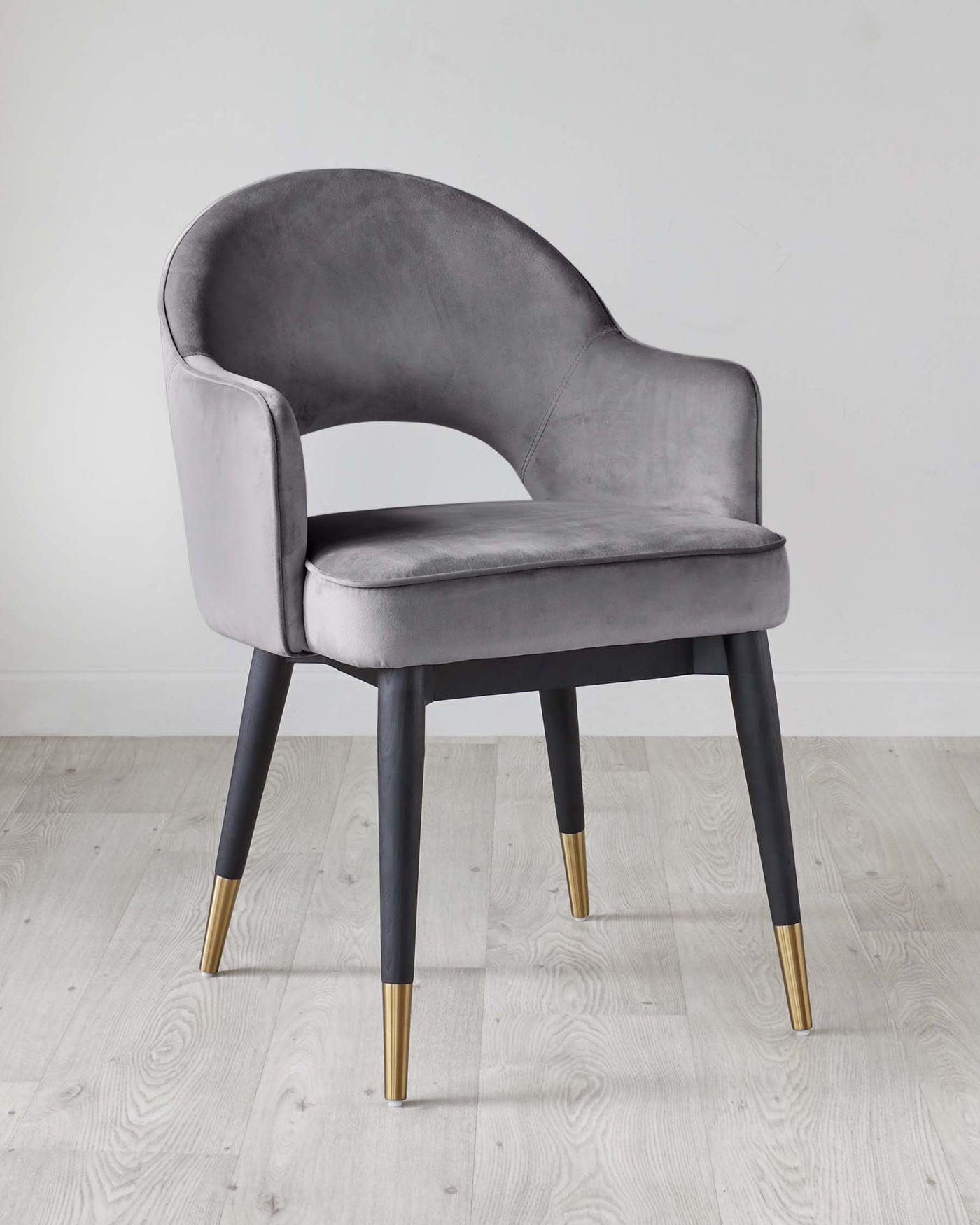 Elegant modern dining chair with velvet upholstery in a soft grey finish, featuring a curved backrest, armless design, cushioned seat, and four tapered legs with contrasting black and gold tips, set against a simplistic white background with a light wood floor.