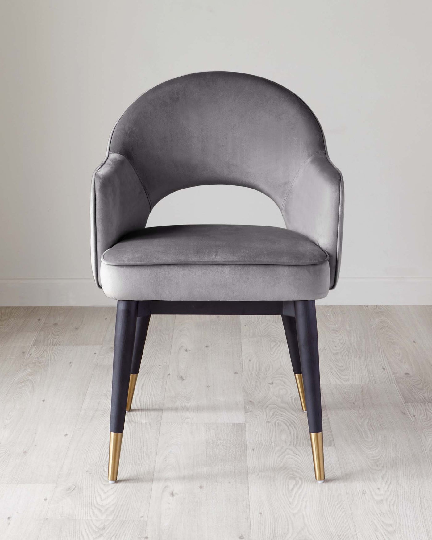 Elegant modern chair with a curved backrest and plush grey velvet upholstery, featuring black tapered legs with gold-toned tips, against a light wooden floor and a pale background.