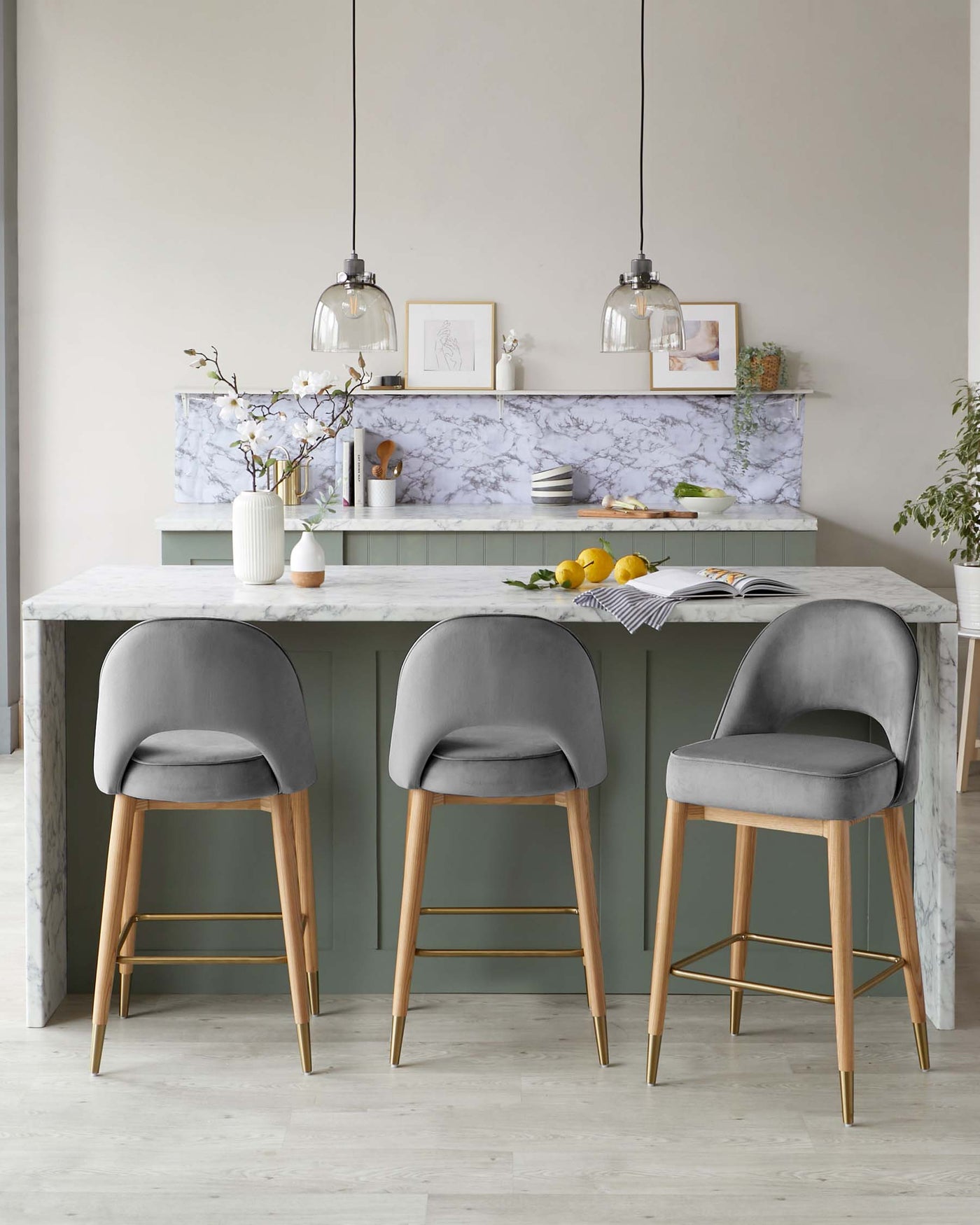 Three modern grey upholstered bar stools with backrest and wooden legs accented with gold tips are positioned at a kitchen island with a marble countertop.
