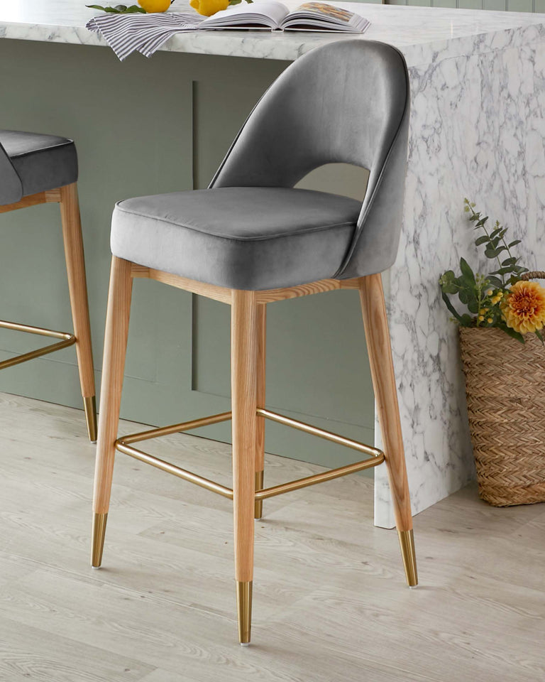 Elegant modern bar stool with grey velvet upholstery, featuring a curved backrest and a cushioned seat. The chair has a solid wood frame with natural finish, accentuated with gold-toned metal footrest and leg tips, adding a touch of sophistication.