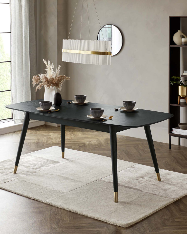A contemporary black dining table with tapered legs finished in brass caps, set on a cream geometric-patterned area rug. The table is accessorized with dark-coloured bowls and plates, complemented by a sleek vase holding pampas grass. Above the table hangs a modern white pendant light with gold accents.
