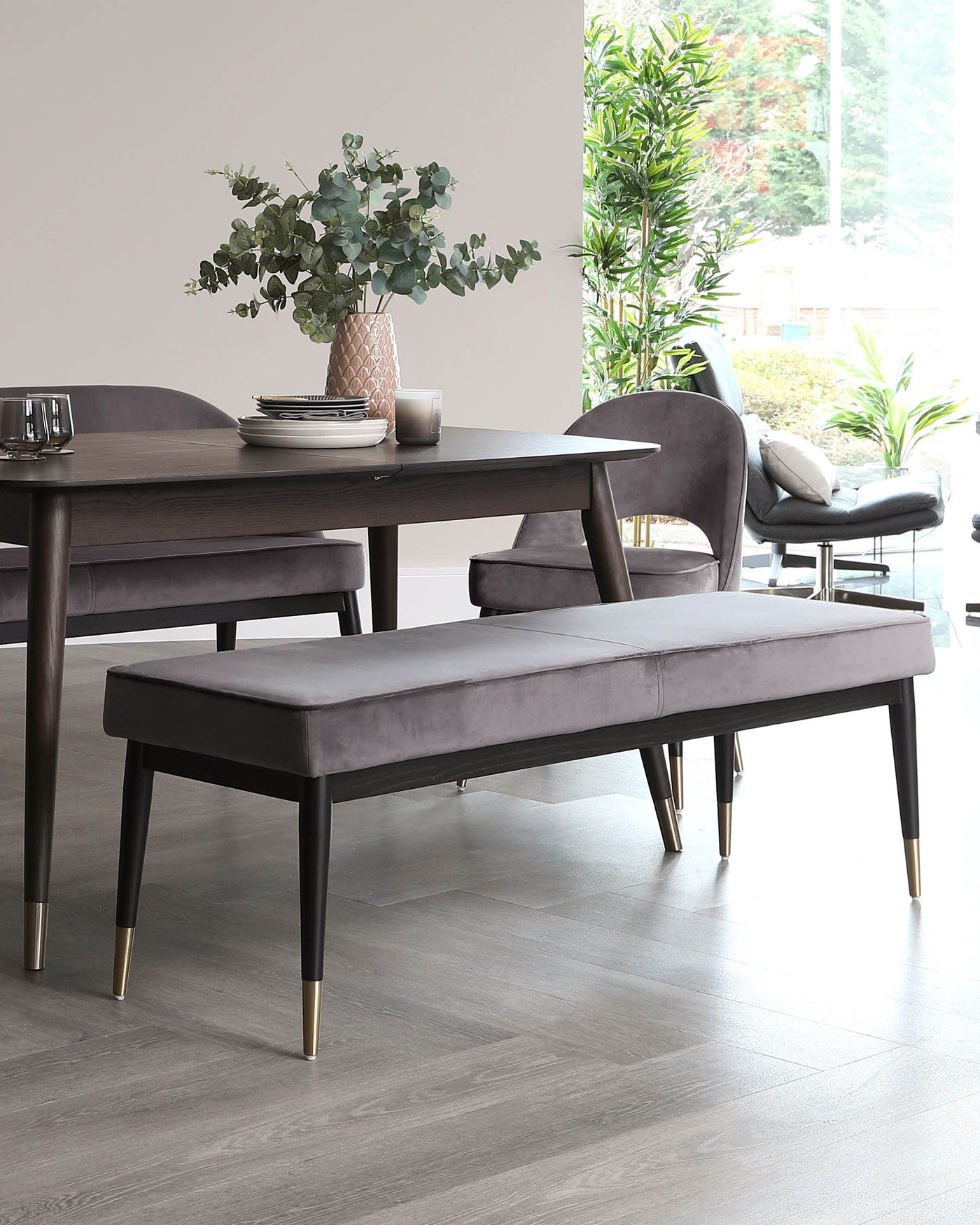 Modern dining room furniture featuring a dark wood table with a matte finish, surrounded by upholstered chairs with curved backs and a matching bench with gold-toned metal capped legs. The setting is complemented by indoor plants and minimalist decor.