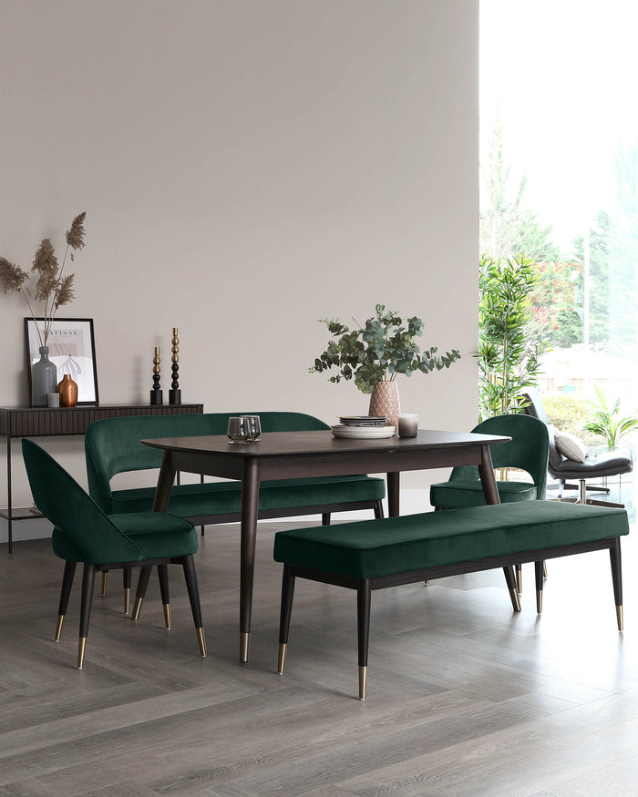 Modern dining room set with a rectangular dark wooden table, surrounded by four green velvet upholstered chairs with black legs tipped in gold, and a matching green velvet bench. The setting reflects a contemporary design in a minimalist interior.