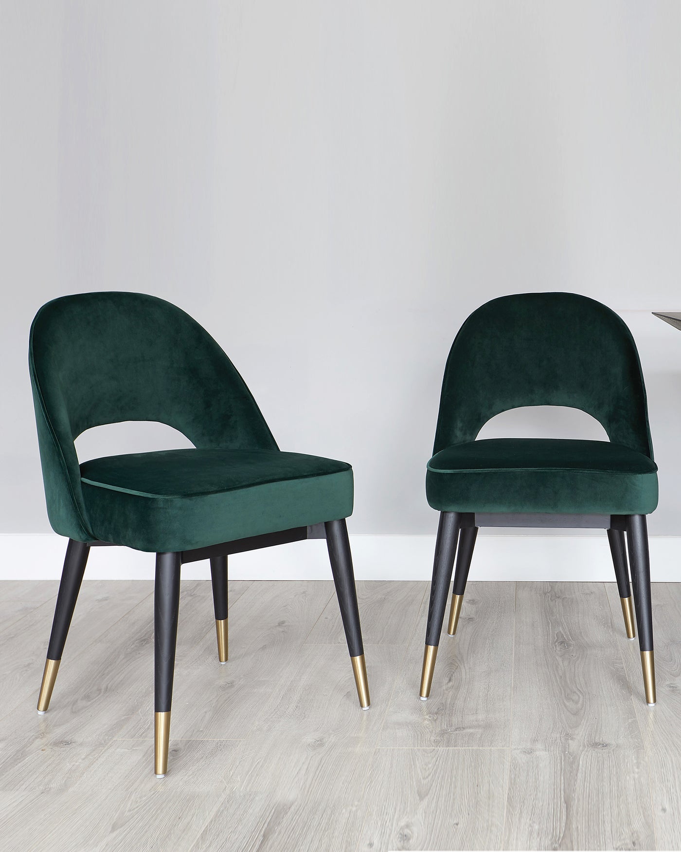 Two modern-style velvet dining chairs in dark green with a curved backrest design and cut-out. The chairs have angled black wooden legs with gold metal tips. The setting is a simple room with light wooden flooring and a plain white wall in the background.