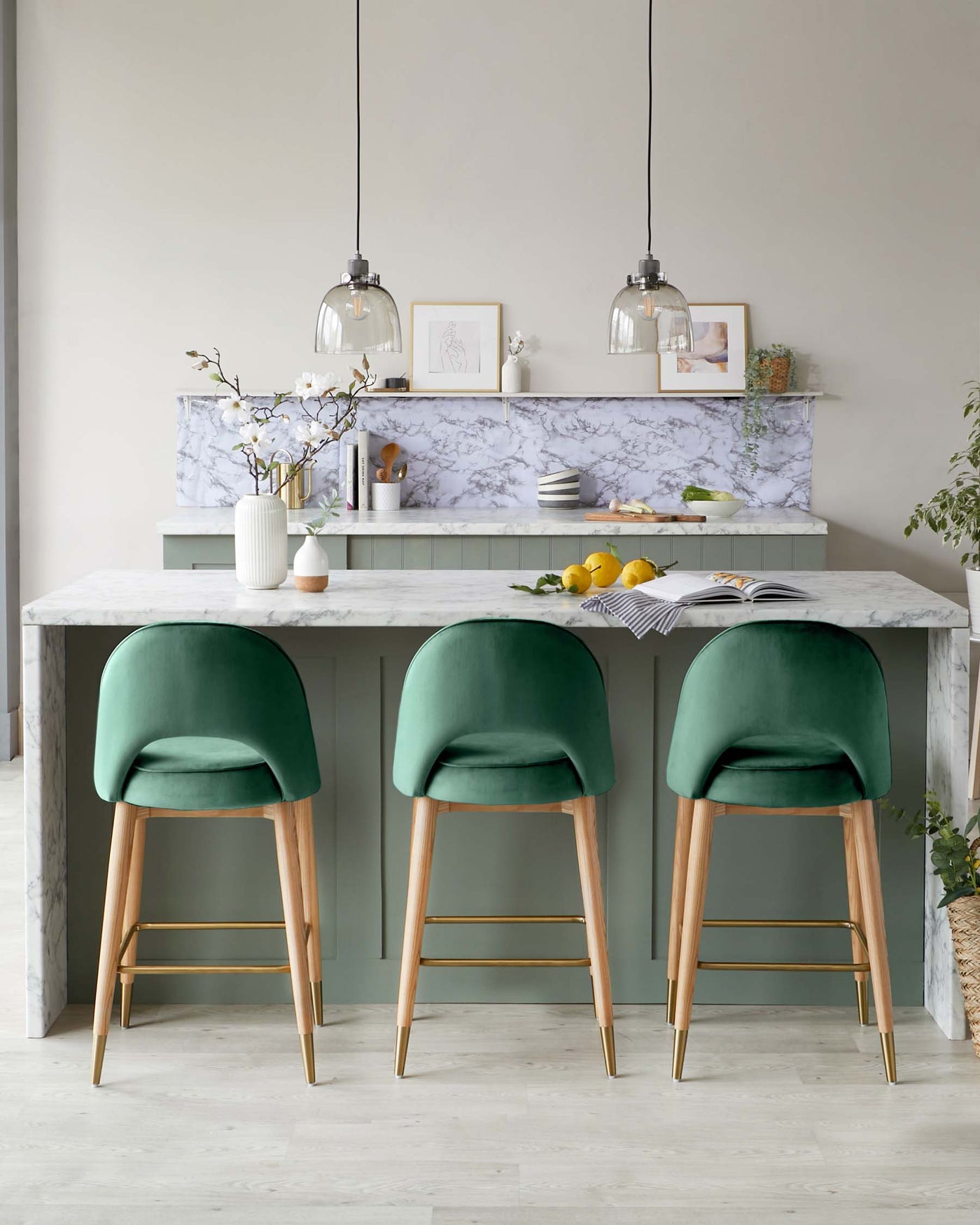 Three modern velvet-upholstered bar stools in rich green with slim wooden legs tipped with gold accents, aligned in front of a marble-topped kitchen island with cabinet storage.