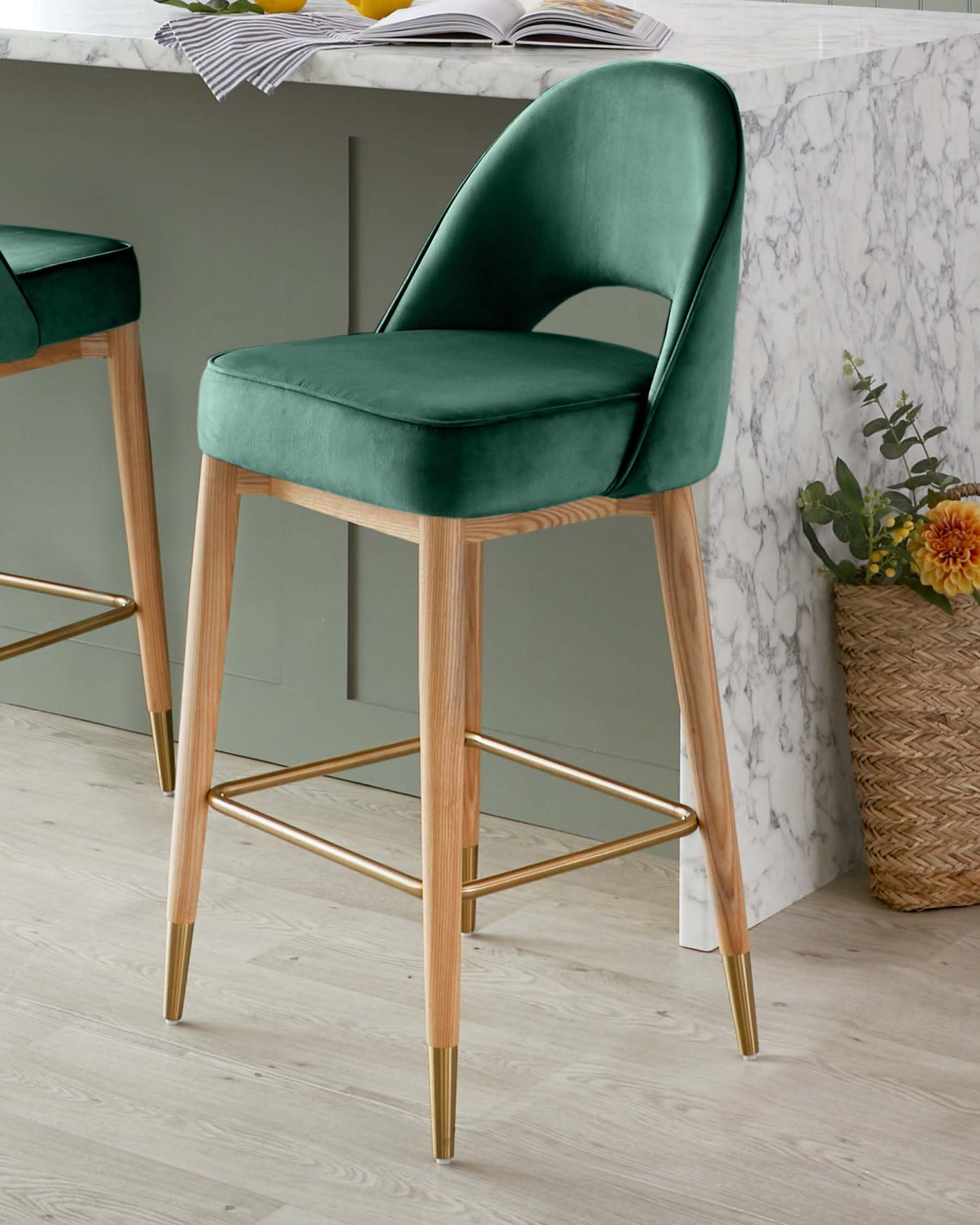 Elegant modern bar stool with a plush, emerald green velvet seat and backrest, set atop a sleek wooden frame with light wood finish and accented with gold-tone footrests and leg caps.