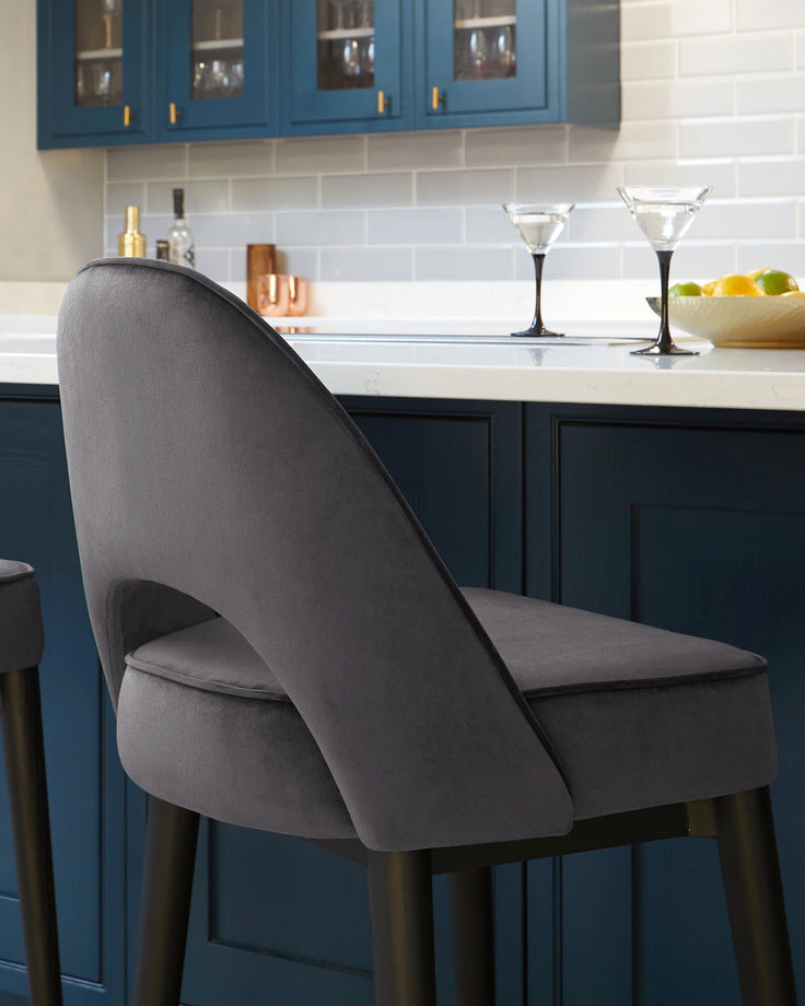 A modern grey upholstered bar stool with a curved backrest and cushioned seat, featuring sleek black legs that taper towards the bottom and are tipped with gold-coloured accents.