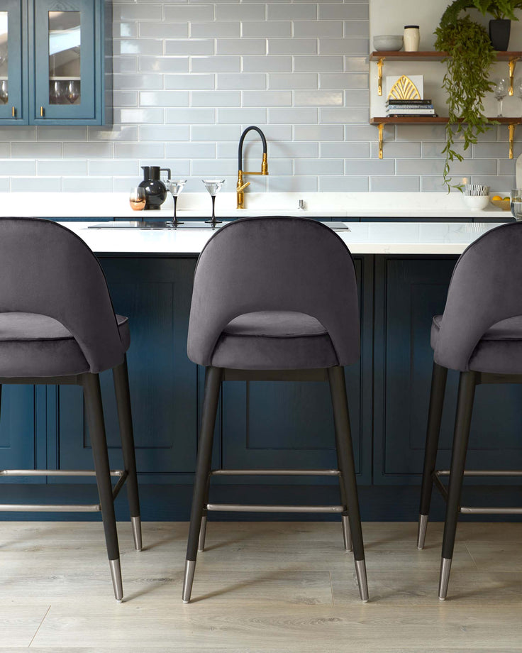 Three contemporary upholstered bar stools with dark grey fabric and slender metallic legs, positioned at a kitchen island with a white countertop.