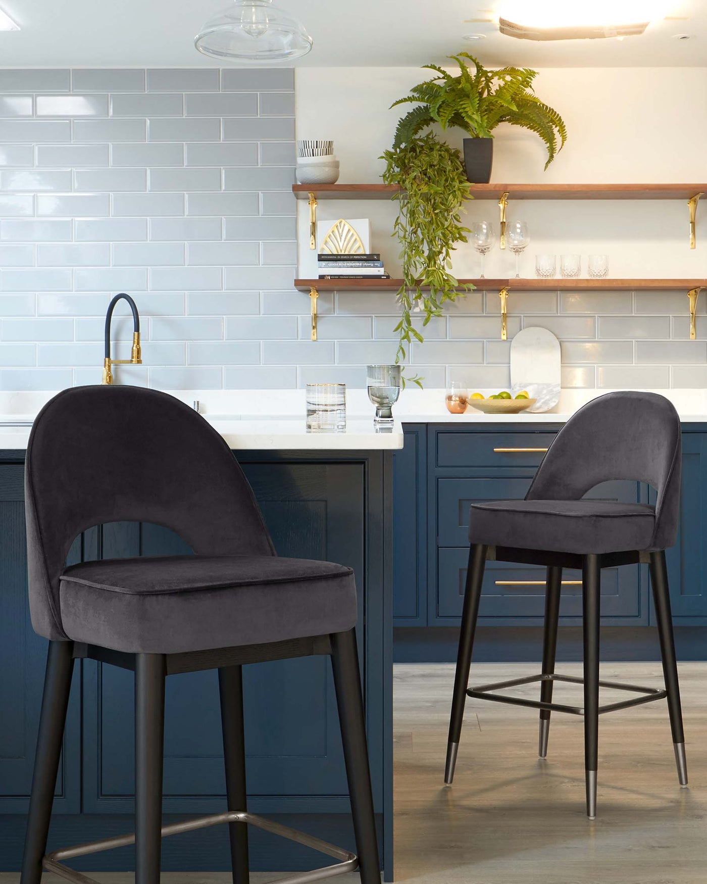 Elegant pair of upholstered bar stools with plush, curved backrests and cushioned seats in a rich, dark grey velvet fabric. The stools stand on sleek, black metal legs with a subtle flare and incorporate a circular, all-around footrest for added comfort. The sophisticated design aligns with a modern kitchen interior, complementing the deep blue cabinetry and warm wooden accents.