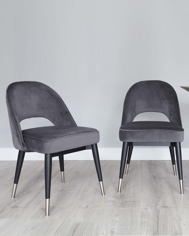 Two contemporary-style dining chairs with plush grey velvet upholstery and contrasting black legs with gold metal tips.