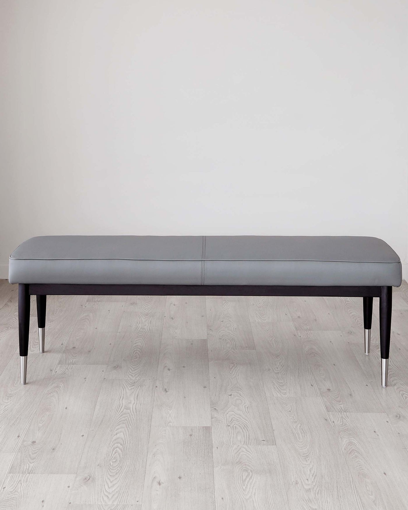 Modern minimalist grey upholstered bench with black wooden legs and metallic accents, displayed on a light wooden floor against a plain white wall.
