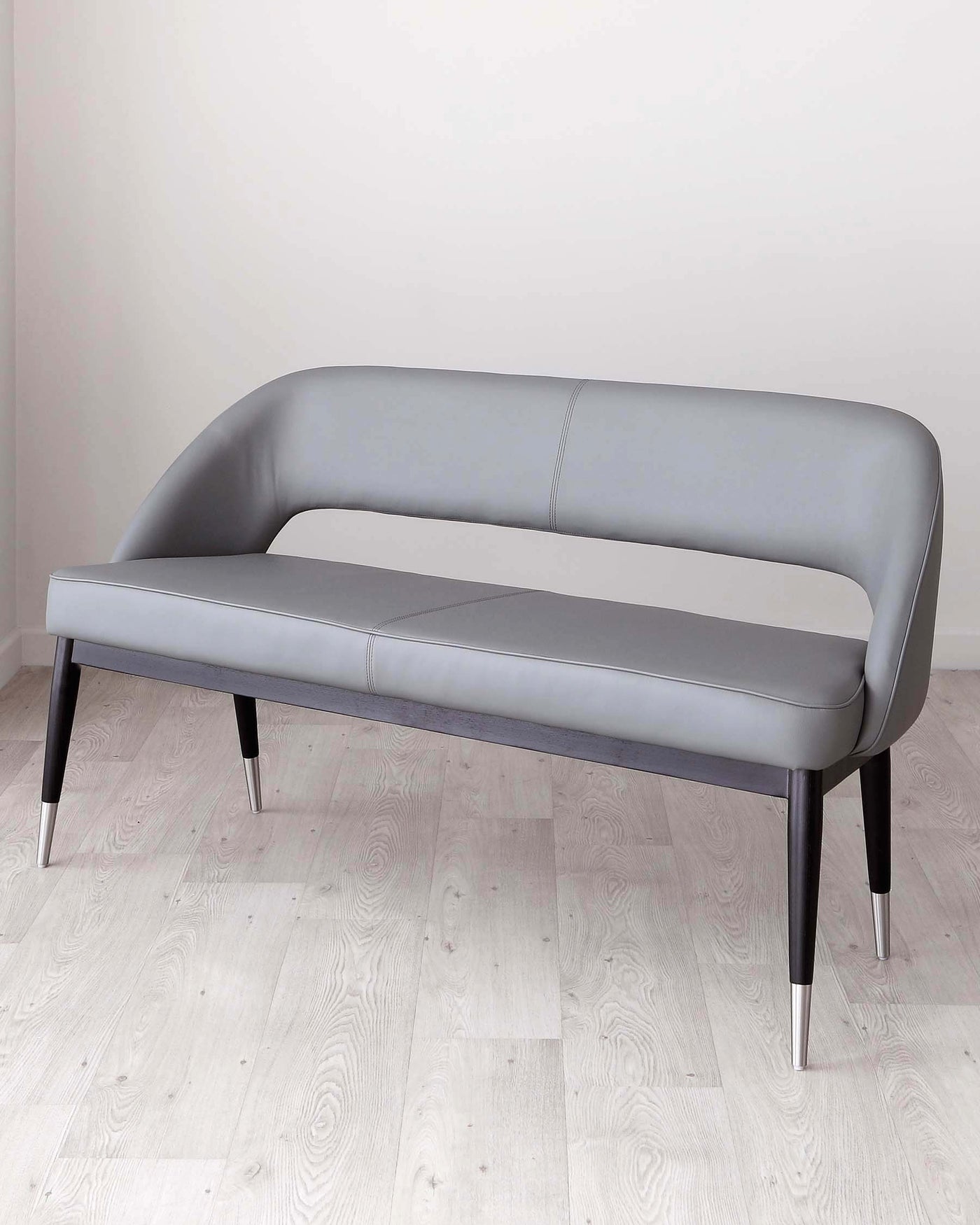 Modern minimalist two-seater sofa with a sleek, curved silhouette, upholstered in a light grey, leather-like material with subtle horizontal stitching. The sofa features a low back and arms, gently arched for a fluid, contemporary design. It stands on four tapered legs, the front two capped with metallic accents, complementing its sophisticated and chic aesthetic.