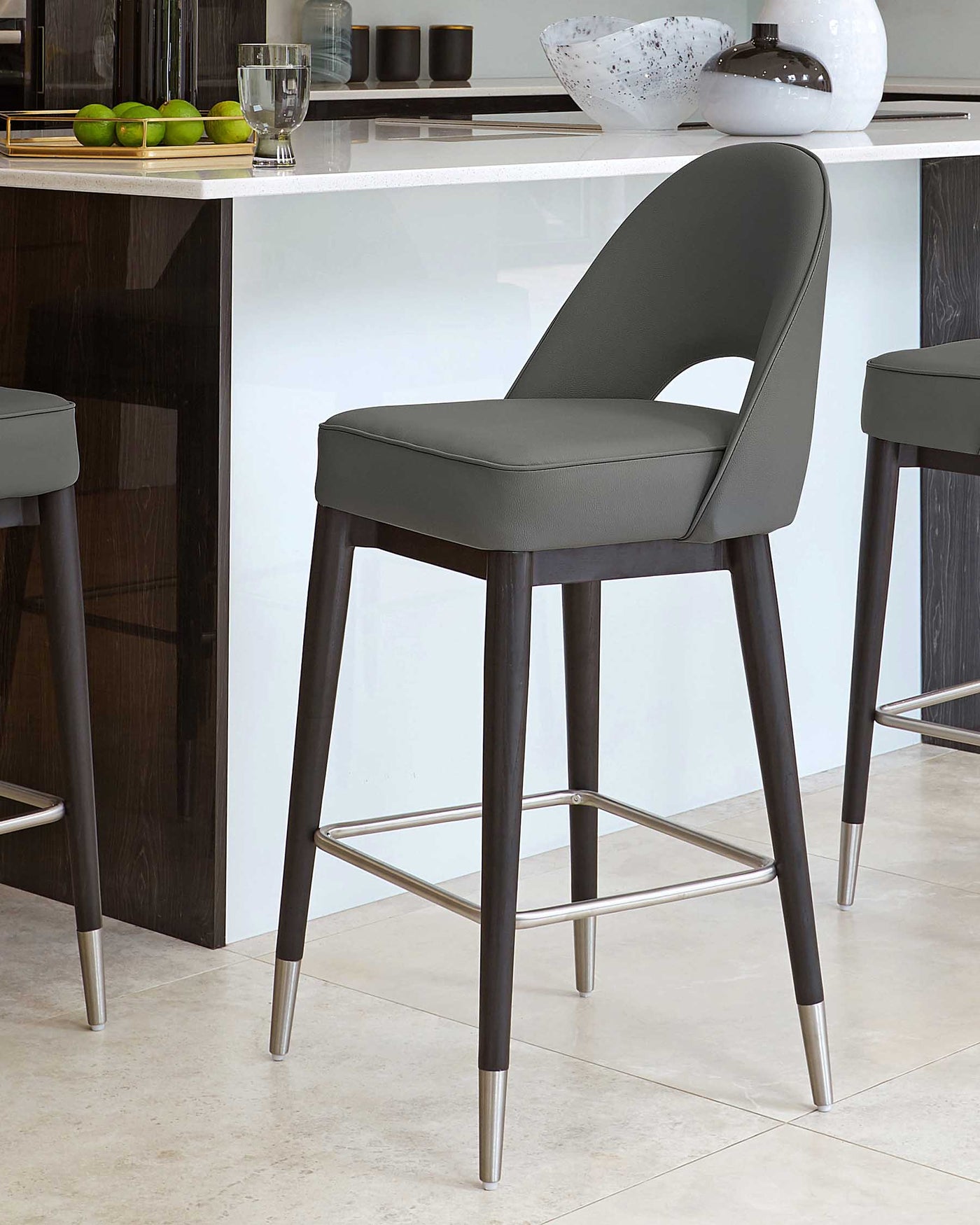Modern charcoal grey bar stool with sleek curves, a rounded seat back, and slim, angled wood legs tipped with metallic accents, featuring a footrest for comfort.