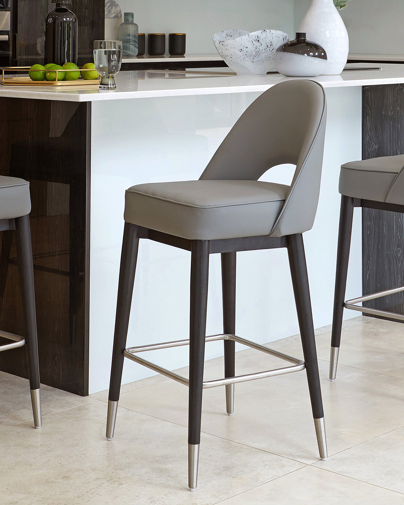 Modern bar stool with a curved leatherette seat in a muted grey tone, featuring slender black legs tipped with metallic accents and a circular footrest.