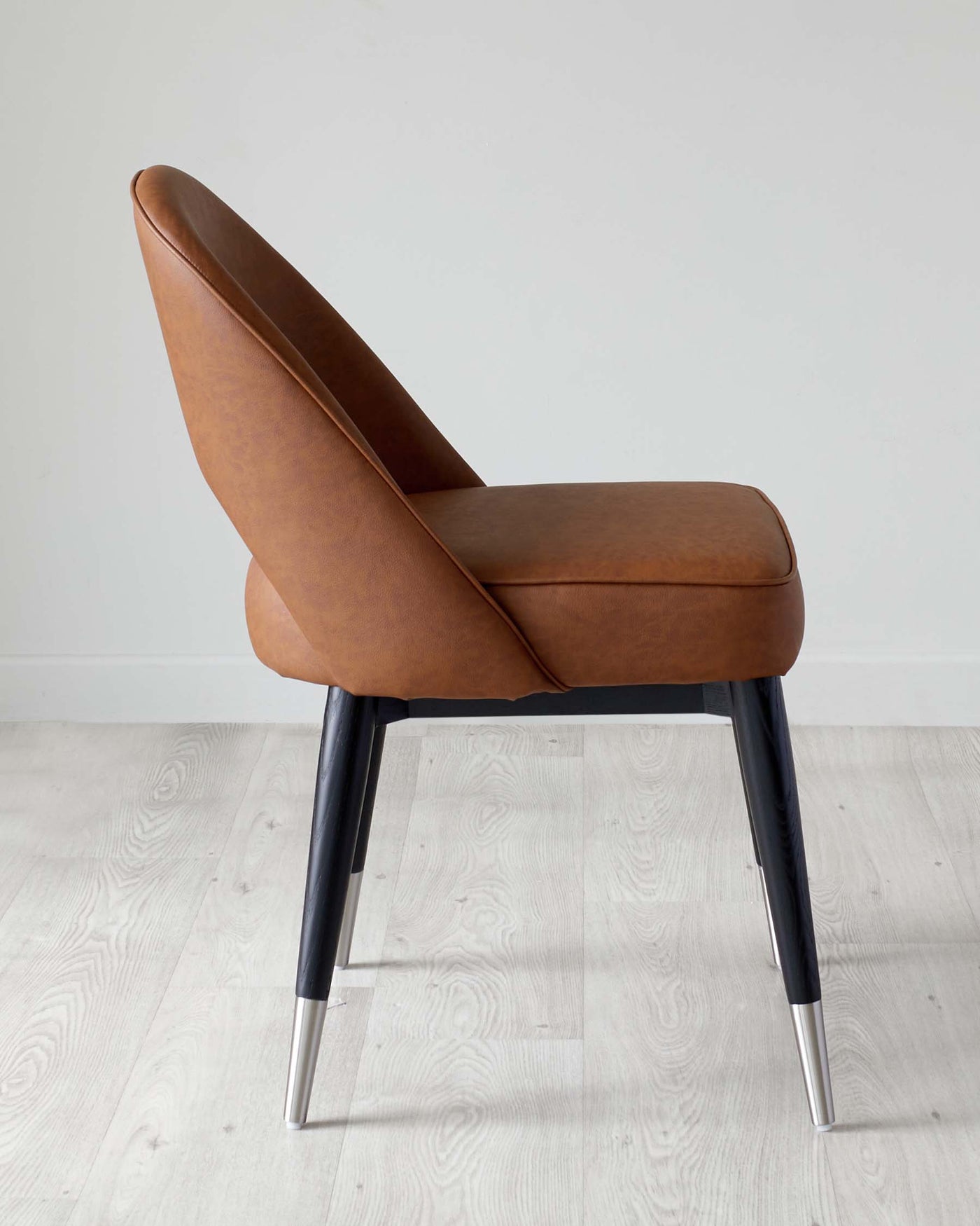 Modern chair with smooth camel-coloured leather upholstery and metal legs, two of which are visible, tapered with a glossy chrome finish. The chair has a curved backrest and a minimalist design, set against a light wooden floor and a white background.