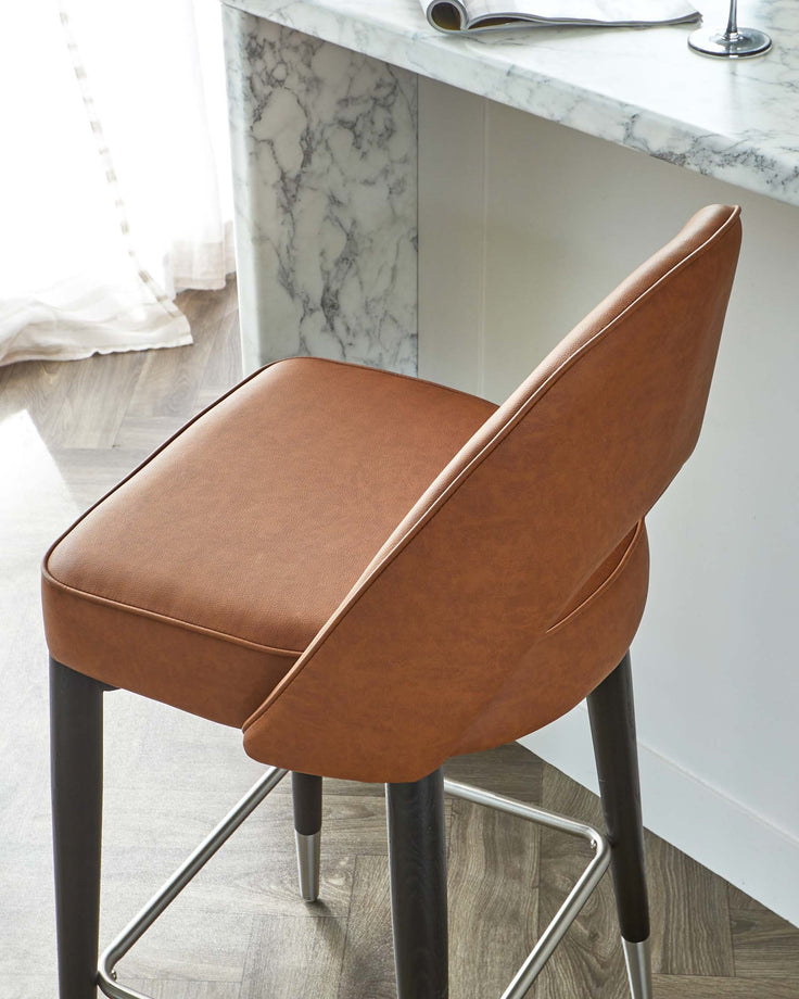 Elegant modern bar stool with a sleek, caramel-brown faux leather seat and a sturdy metal frame in black, featuring slim, tapered legs with metallic foot rests. Positioned next to a marble countertop.