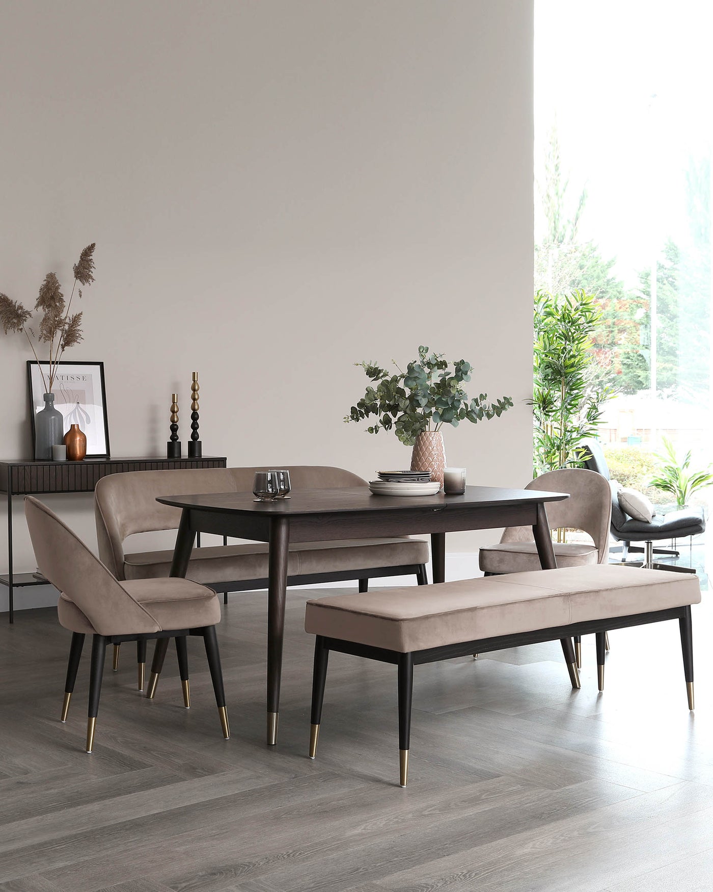 Contemporary dining room featuring a dark wooden table with a smooth finish, flanked by elegant taupe upholstered chairs with black legs tipped with gold accents. A matching bench with the same fabric and design completes the set. A sleek sideboard in the background ties together the modern aesthetic.