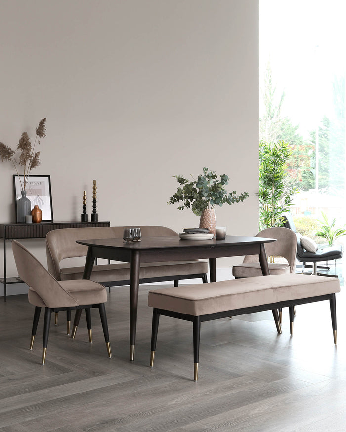 Contemporary dining room featuring a dark wooden table with a smooth finish, flanked by elegant taupe upholstered chairs with black legs tipped with gold accents. A matching bench with the same fabric and design completes the set. A sleek sideboard in the background ties together the modern aesthetic.